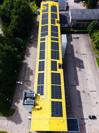 Aerial view of a long yellow industrial building covered with solar panels surrounded by a paved parking lot and green trees in Valdlauci Kekava Municipality Latvia. Aerial view of a long yellow industrial building covered with solar panels surrounded by a paved parking lot and green trees in Valdlauci Kekava Municipality Latvia.