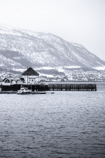 View of a waterfront building and wooden pier with snow covered mountains in the background across the water in Tromso Troms Norway. View of a waterfront building and wooden pier with snow covered mountains in the background across the water in Tromso Troms Norway.