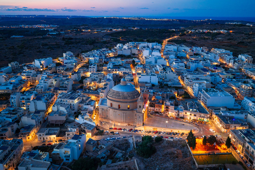 Aerial view of the Mgarr Parish Church illuminated at night surrounded by the dense urban fabric of the town with warm street lights glowing in Mgarr Northern Region Malta.