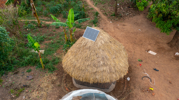 Aerial view of a traditional thatched roof hut with a modern solar panel installed on top surrounded by dirt paths and banana trees in Mbale Eastern Region Uganda. Aerial view of a traditional thatched roof hut with a modern solar panel installed on top surrounded by dirt paths and banana trees in Mbale Eastern Region Uganda.