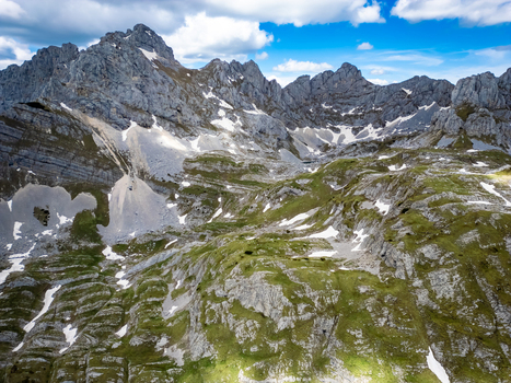 Aerial view of Bobotov Kuk in the Durmitor mountains featuring rugged rocky peaks patches of snow and green slopes under a bright blue sky in Zabljak Municipality Montenegro. Aerial view of Bobotov Kuk in the Durmitor mountains featuring rugged rocky peaks patches of snow and green slopes under a bright blue sky in Zabljak Municipality Montenegro.