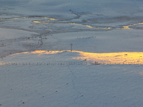 Aerial view of the golden light kissing the snow covered hills and a cross with a winding river snaking through the landscape Nasbinals Occitanie France.