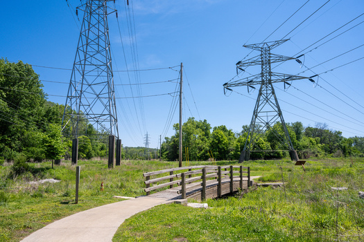 View of a wooden bridge on a paved walking path in Greentree Park with high voltage power line towers and lush green trees under a clear blue sky in Kirkwood Missouri United States. View of a wooden bridge on a paved walking path in Greentree Park with high voltage power line towers and lush green trees under a clear blue sky in Kirkwood Missouri United States.