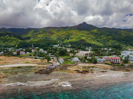 Aerial view of waves crashing against the rocky shore beneath verdant hills dotted with buildings framed by a cloudy sky Victoria Seychelles. Aerial view of waves crashing against the rocky shore beneath verdant hills dotted with buildings framed by a cloudy sky Victoria Seychelles.