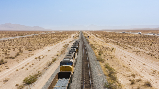 Aerial view of a freight train traveling along tracks through a vast desert landscape with a highway in the distance in the United States.