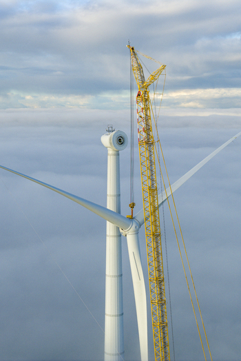 Aerial view of a wind turbine under construction with a large yellow crane above a thick layer of clouds under a soft blue sky.