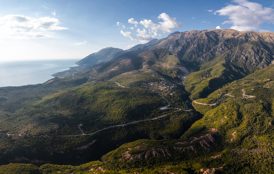 Aerial view of Gjipe Canyon featuring rugged mountain terrain lush green vegetation winding roads and the coastline of the Ionian Sea in Iliaz Vlore County Albania. Aerial view of Gjipe Canyon featuring rugged mountain terrain lush green vegetation winding roads and the coastline of the Ionian Sea in Iliaz Vlore County Albania.