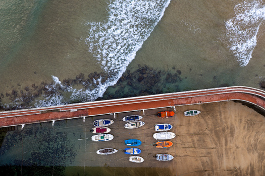Aerial view of Puerto de Zarautz with small boats resting on the sandy shore beside a long pier and waves breaking against the rocks in Zarautz Basque Country Spain.