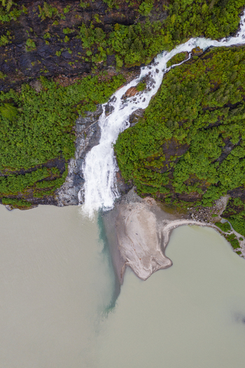 Aerial view of a cascading waterfall flowing into a glacial lake surrounded by lush green forest and rocky terrain in Juneau Alaska United States. Aerial view of a cascading waterfall flowing into a glacial lake surrounded by lush green forest and rocky terrain in Juneau Alaska United States.