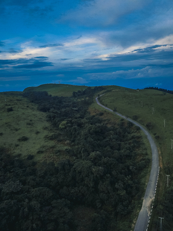 Aerial view of a winding grey road cutting through vibrant green hills and dense dark forest under a dramatic blue sky Obudu Cross River Nigeria.
