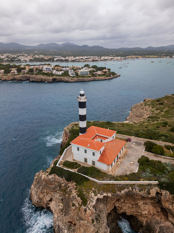 Aerial view of the Portocolom Lighthouse on a rocky cliff overlooking the turquoise sea and coastal town under a cloudy sky in Portocolom Balearic Islands Spain. Aerial view of the Portocolom Lighthouse on a rocky cliff overlooking the turquoise sea and coastal town under a cloudy sky in Portocolom Balearic Islands Spain.