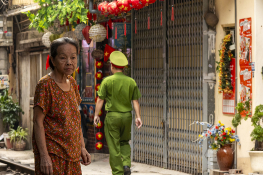 Hanoi Vietnam 11 November 2025 View of an elderly woman in a narrow street with a police officer walking in the background near traditional lanterns flags and potted plants.