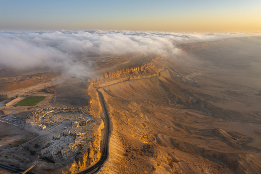 Aerial view of the Mitzpe Ramon crater at sunset with low clouds rolling over the desert cliffs a winding road and buildings in Mitzpe Ramon South District Israel. Aerial view of the Mitzpe Ramon crater at sunset with low clouds rolling over the desert cliffs a winding road and buildings in Mitzpe Ramon South District Israel.