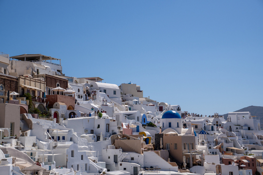 View of whitewashed houses and a blue domed church on the steep cliffs of Santorini with people walking along the paths under a bright sky Fira Greece. View of whitewashed houses and a blue domed church on the steep cliffs of Santorini with people walking along the paths under a bright sky Fira Greece.