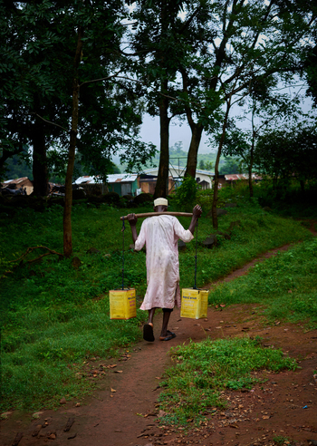 Riyom Plateau Nigeria 17 September 2026 View of a man carrying two yellow water containers on a wooden yoke walking along a dirt path through a lush green landscape.