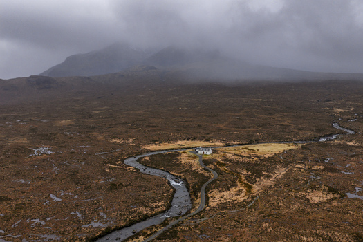 Aerial view of a lonely white cottage nestled by a winding river in the vast moorland landscape under misty mountains Isle of Skye Scotland United Kingdom. Aerial view of a lonely white cottage nestled by a winding river in the vast moorland landscape under misty mountains Isle of Skye Scotland United Kingdom.