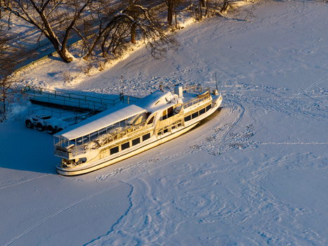 Aerial view of a white passenger boat docked at a pier on a frozen snow covered lake with long shadows from nearby trees in Trakai Vilnius County Lithuania. Aerial view of a white passenger boat docked at a pier on a frozen snow covered lake with long shadows from nearby trees in Trakai Vilnius County Lithuania.