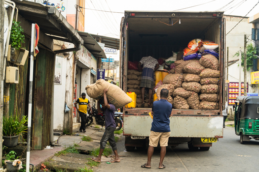 Sri Lanka 15 February 2026 View of workers unloading heavy sacks of produce from a large truck on a busy street with a green tuk tuk passing by.