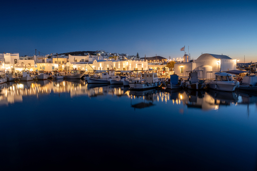View of fishing boats moored in the harbor with illuminated white buildings and reflections on the calm water at night in Lefkada Greece.