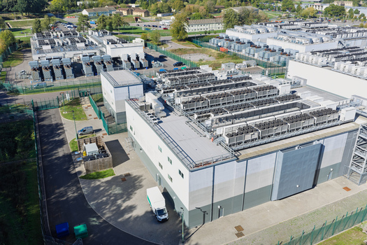 Aerial view of a large data center facility featuring rows of cooling units on the roof surrounding paved areas and security fencing in the United Kingdom.