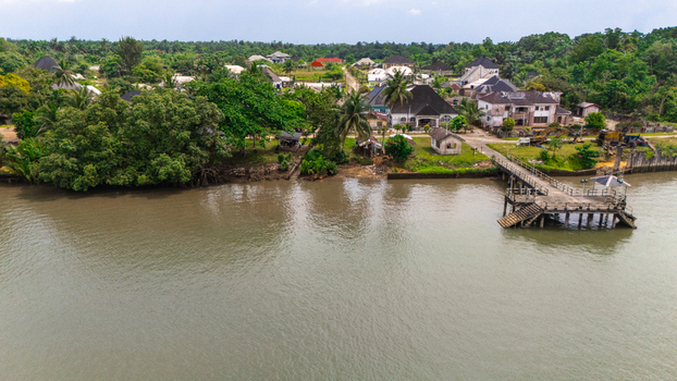 Aerial view of houses and lush trees along the riverbank with a small concrete pier extending into the water in Minima Rivers Nigeria.