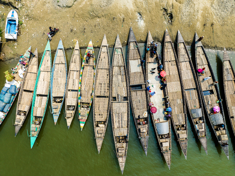 Aerial view of traditional wooden boats docked along the riverbank with people moving between the vessels and cargo loaded on the decks in Tangail Dhaka Division Bangladesh. Aerial view of traditional wooden boats docked along the riverbank with people moving between the vessels and cargo loaded on the decks in Tangail Dhaka Division Bangladesh.