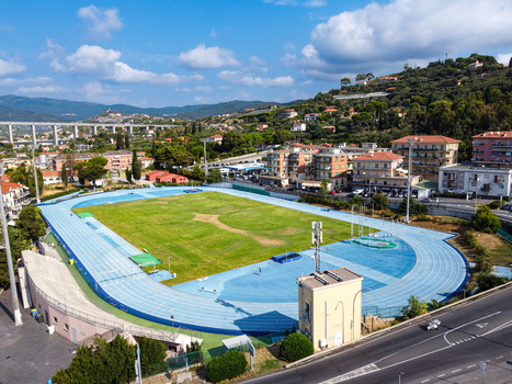 Aerial view of an athletics stadium with a blue running track and green grass field surrounded by urban buildings and lush hills Imperia Liguria Italy. Aerial view of an athletics stadium with a blue running track and green grass field surrounded by urban buildings and lush hills Imperia Liguria Italy.
