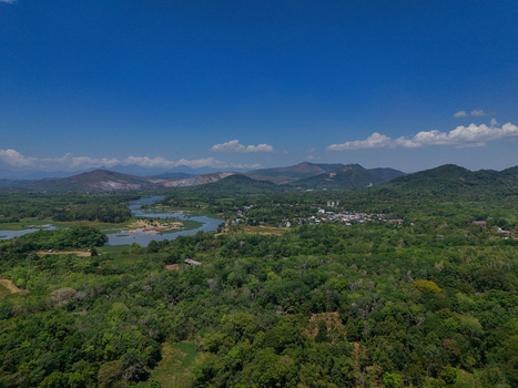 Aerial view of a winding river flowing through a lush green landscape with dense forests a small village and distant mountains under a clear blue sky in Indonesia. Aerial view of a winding river flowing through a lush green landscape with dense forests a small village and distant mountains under a clear blue sky in Indonesia.