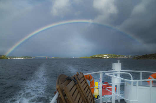 View of a vibrant rainbow arching over the sea with the Skye Bridge in the distance seen from the deck of a boat under a dramatic stormy sky in Isle of Skye United Kingdom. View of a vibrant rainbow arching over the sea with the Skye Bridge in the distance seen from the deck of a boat under a dramatic stormy sky in Isle of Skye United Kingdom.