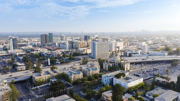 Aerial view of the Los Angeles cityscape featuring modern office buildings residential areas and a major highway interchange under a clear blue sky in Los Angeles California United States.