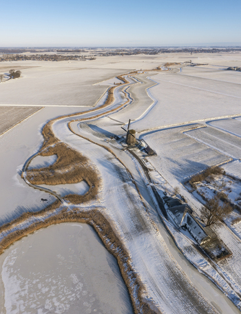 Aerial view of a traditional Dutch windmill standing beside a frozen winding canal in a snow covered landscape in Ursem North Holland Netherlands.
