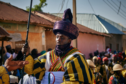 Zaria city Nigeria 21 March 2026 View of a regal figure in a vibrant striped robe and majestic purple turban holding a staff on a sunlit street. Zaria city Nigeria 21 March 2026 View of a regal figure in a vibrant striped robe and majestic purple turban holding a staff on a sunlit street.
