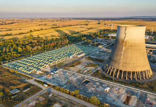 Aerial view of an industrial power site featuring a large cooling tower and a battery storage facility surrounded by agricultural fields in warm evening light Italy.
