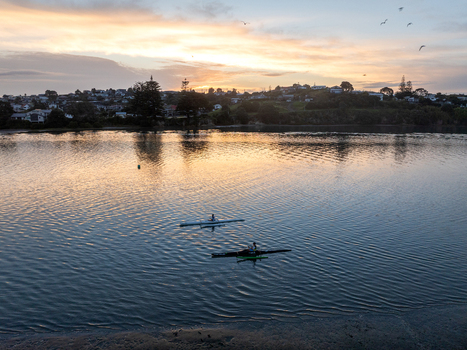 Raglan New Zealand 09 February 2026 Aerial view of two people kayaking on calm water at sunset with the town of Raglan in the background under a warm glowing sky.