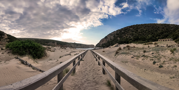 View of Spiaggia di Cala Domestica with a wooden walkway leading through sand dunes and green hills under a cloudy sky Provincia del Sulcis Iglesiente Sardegna Italy. View of Spiaggia di Cala Domestica with a wooden walkway leading through sand dunes and green hills under a cloudy sky Provincia del Sulcis Iglesiente Sardegna Italy.