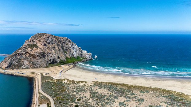 Aerial view of Morro Rock standing prominently beside a sandy beach and the blue Pacific Ocean under a clear sky in Morro Bay California United States.