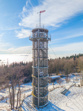 Aerial view of Majak Jary Cimrmana observation tower surrounded by a snowy forest landscape under a clear blue sky in Korenov Liberecky kraj Czechia. Aerial view of Majak Jary Cimrmana observation tower surrounded by a snowy forest landscape under a clear blue sky in Korenov Liberecky kraj Czechia.