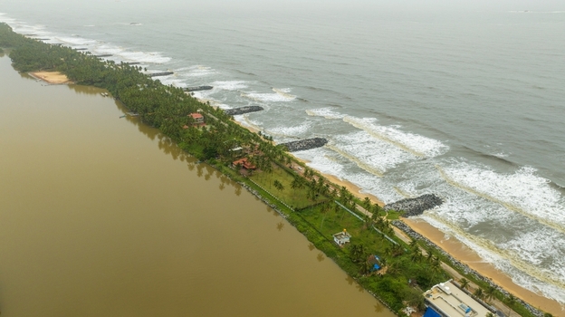 Aerial view of Mattu Beach with its narrow strip of land separating the backwaters from the Arabian Sea featuring coconut palms and rocky breakwaters in Udupi Karnataka India.