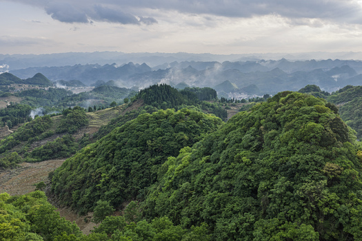 Aerial view of lush green karst mountains and rolling hills under a cloudy sky with mist in the valleys Gaoxing Village Guizhou China.