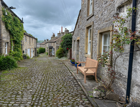 View of a narrow cobblestone street lined with historic stone cottages and a wooden bench under an overcast sky in the Yorkshire Dales Hawes England United Kingdom. View of a narrow cobblestone street lined with historic stone cottages and a wooden bench under an overcast sky in the Yorkshire Dales Hawes England United Kingdom.