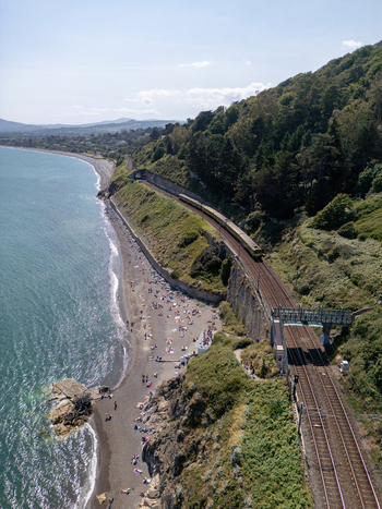 Aerial view of a train on coastal tracks near Dalkey with a beach filled with people lush green hills and the blue sea in Dublin County Dublin Ireland. Aerial view of a train on coastal tracks near Dalkey with a beach filled with people lush green hills and the blue sea in Dublin County Dublin Ireland.