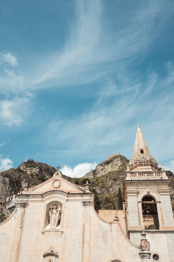 View of a cream colored church facade with a steeple reaching toward a wispy blue sky backed by rugged cliffs in Taormina Catania Italy.