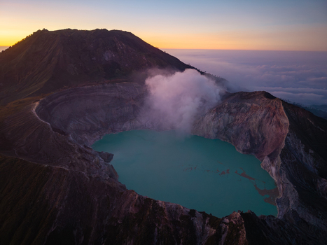 Aerial view of the turquoise crater lake at Ijen volcano with rising sulfur smoke and rugged volcanic slopes during golden hour in Ijen East Java Indonesia.