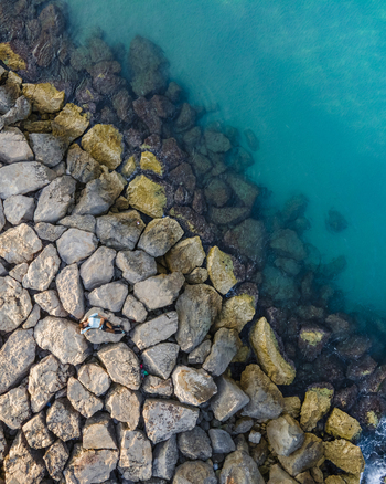 Aerial view of a person lying on large rocks of a breakwater by the turquoise sea water in Sitges Catalonia Spain. Aerial view of a person lying on large rocks of a breakwater by the turquoise sea water in Sitges Catalonia Spain.
