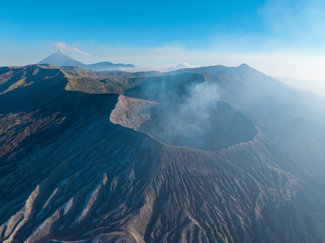 Aerial view of Mount Bromo crater with volcanic smoke rugged terrain and surrounding mountain peaks under a clear blue sky in Tosari East Java Indonesia.