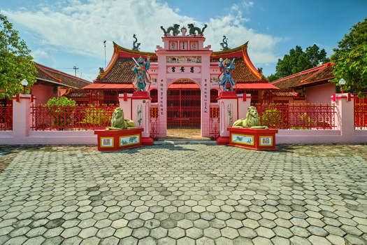 View of Tjoe An Kiong Temple featuring an ornate pink facade traditional statues and a stone courtyard under a clear sky in Kecamatan Lasem Jawa Tengah Indonesia.