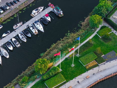 Aerial view of a marina with various boats docked at a pier featuring a green lawn with flags flying along the riverbank in Gdansk Pomeranian Voivodeship Poland. Aerial view of a marina with various boats docked at a pier featuring a green lawn with flags flying along the riverbank in Gdansk Pomeranian Voivodeship Poland.