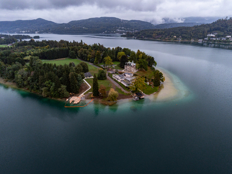 Aerial view of the Maria Worth peninsula on Lake Worthersee featuring historic architecture lush trees and calm turquoise water under a cloudy sky in Maria Worth Carinthia Austria.