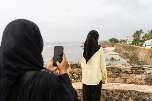 Sri Lanka 07 January 2026 View of two women in hijabs standing on the historic stone walls of Galle Fort while taking a photo of the ocean and coastline with a smartphone.