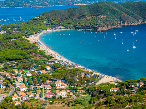 Aerial view of Lacona Beach with its crescent shaped sandy shore turquoise waters anchored sailboats and surrounding pine forests in Lacona Tuscany Italy.